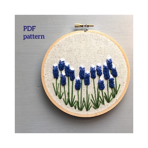 May include: Embroidered hoop art featuring a row of blue and white flowers with green stems on a beige linen background. The hoop is made of wood and has a diameter of approximately 8 inches. The text "PDF pattern" is visible in the top left corner of the image.