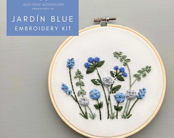 May include: A embroidery kit with a wooden hoop containing a design of blue and gray flowers with green stems. The kit is titled "JARDÍN BLUE EMBROIDERY KIT" and is from "and other adventures EMBROIDERY CO."