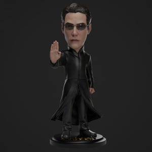 May include: A bobblehead figurine of a man in a long black coat and sunglasses, with his right hand raised in a stopping gesture. The figure is on a black base with gold accents. The figurine is a collectible.