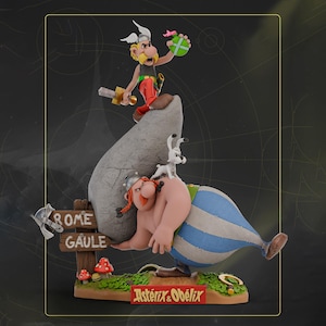 Op de afbeelding: Een gedetailleerd beeldje met de personages Asterix en Obelix. Asterix staat op een rots, met een zwaard en een groen voorwerp. Obelix ligt, met een grote steen. Een kleine witte hond zit op Obelix' rug. De basis heeft de tekst "Rome Gaule" en "Asterix & Obelix".