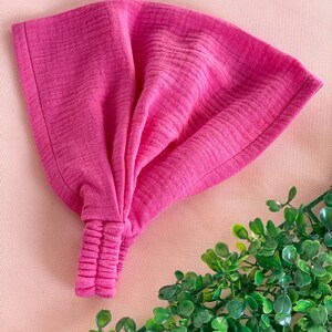 May include: A vibrant pink headband with a textured, crinkled fabric. The headband is knotted at the front and has a gathered elastic section at the back. The accessory is shown on a pink surface with green foliage in the corner.