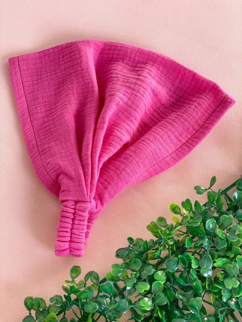 May include: A vibrant pink headband with a textured, crinkled fabric. The headband is knotted at the front and has a gathered elastic section at the back. The accessory is shown on a pink surface with green foliage in the corner.
