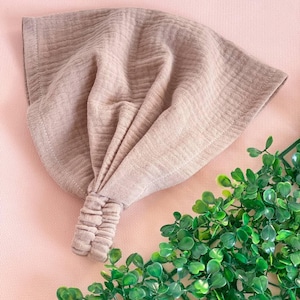 May include: A taupe-colored headband with a knotted design. The fabric has a textured, crinkled appearance. The headband is displayed on a pink surface with green foliage in the corner.