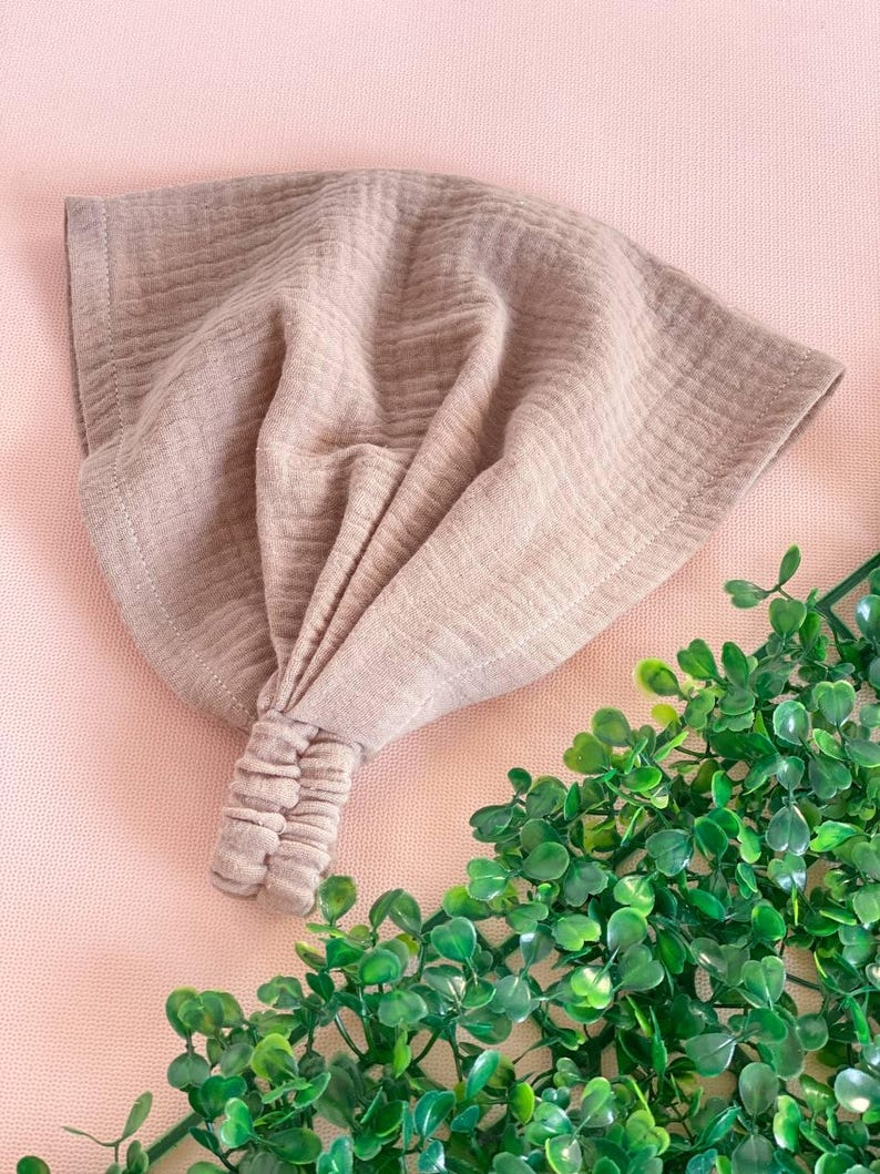 May include: A taupe-colored headband with a knotted design. The fabric has a textured, crinkled appearance. The headband is displayed on a pink surface with green foliage in the corner.