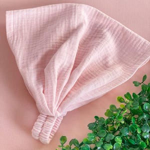 May include: A light pink headband made of a soft, textured fabric. The headband is knotted at the top and has a gathered section at the back. The background is a matching pink color, with a small green plant in the corner.