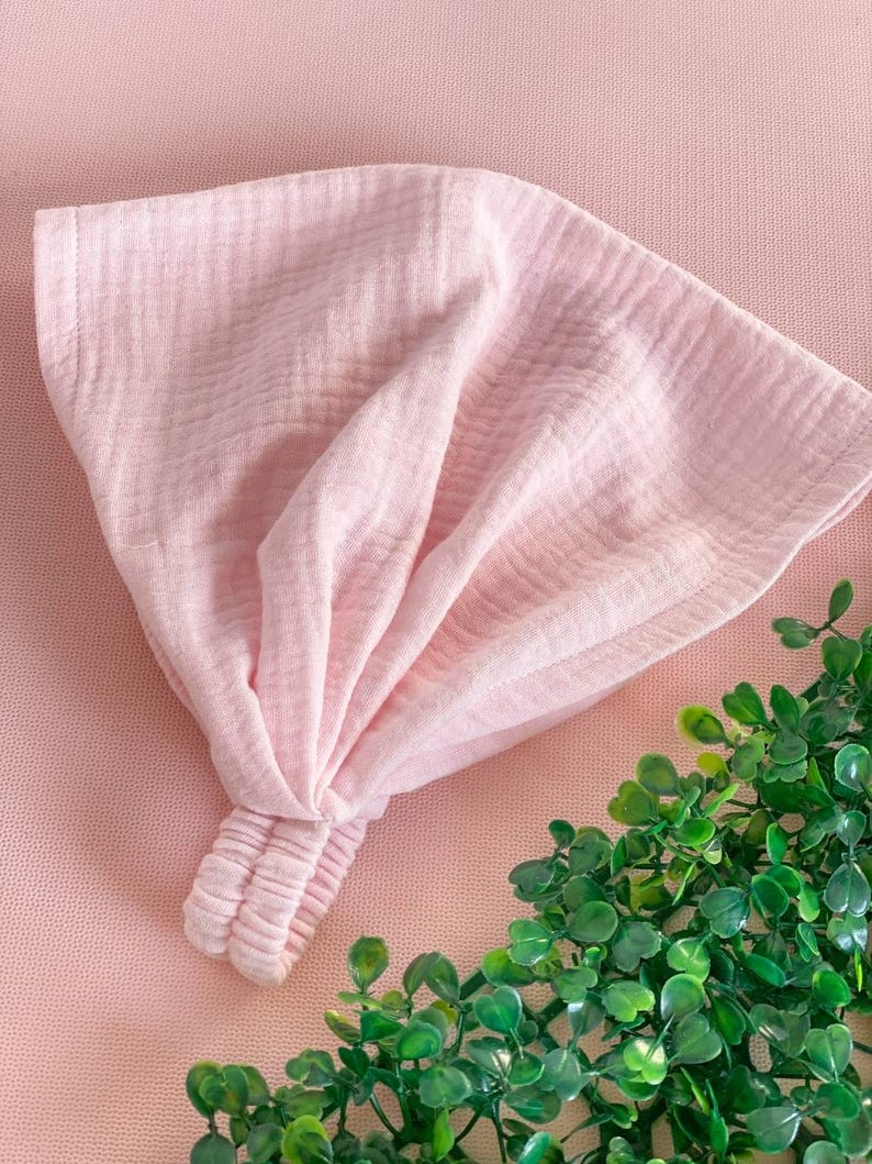 May include: A light pink headband made of a soft, textured fabric. The headband is knotted at the top and has a gathered section at the back. The background is a matching pink color, with a small green plant in the corner.