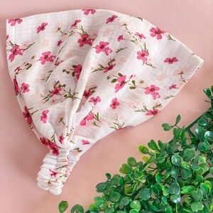 May include: A white headband with a pink floral pattern. The fabric is soft and textured, with small pink flowers and green stems scattered across the surface. The headband is knotted at the top and has an elastic band at the back.