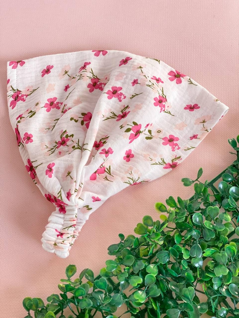 May include: A white headband with a pink floral pattern. The fabric is soft and textured, with small pink flowers and green stems scattered across the surface. The headband is knotted at the top and has an elastic band at the back.