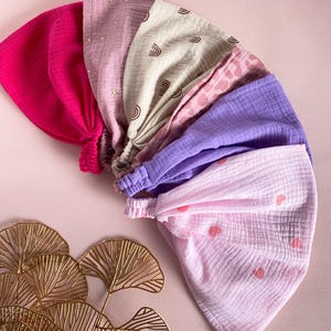 Op de afbeelding: Een verzameling kleurrijke haarbanden in verschillende tinten roze, paars en beige. Sommige haarbanden hebben patronen zoals sterren, regenbogen en harten. De haarbanden zijn in een waaier vorm gerangschikt, waarbij hun verschillende ontwerpen en texturen worden getoond.