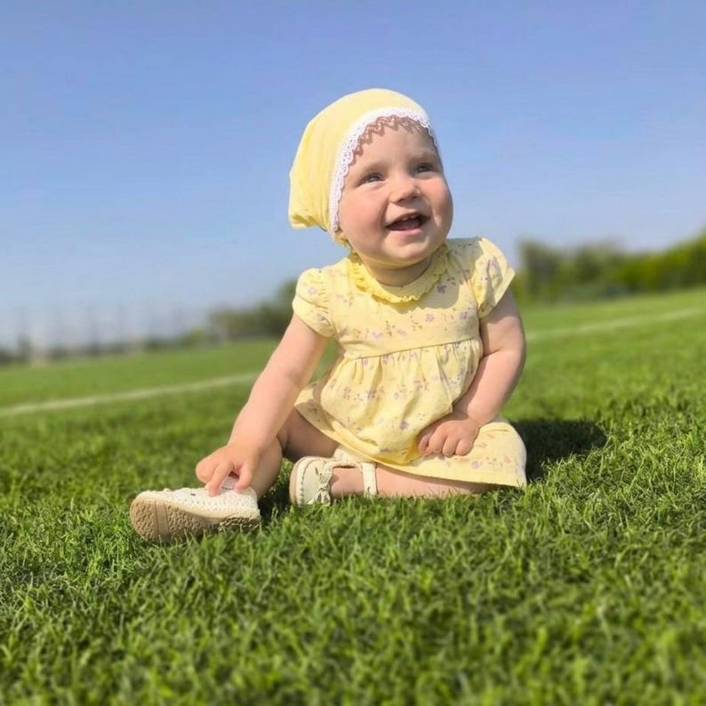 May include: A smiling baby wearing a yellow dress and matching bonnet with white lace trim sits on green grass under a clear blue sky. The baby is also wearing white shoes with a strap.
