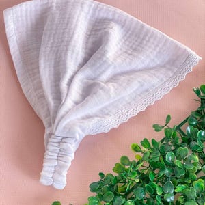 May include: A white headband with a textured fabric and a delicate lace trim. The headband is gathered at the center and appears to be made of a soft, lightweight material. The background is a light pink surface with green foliage.