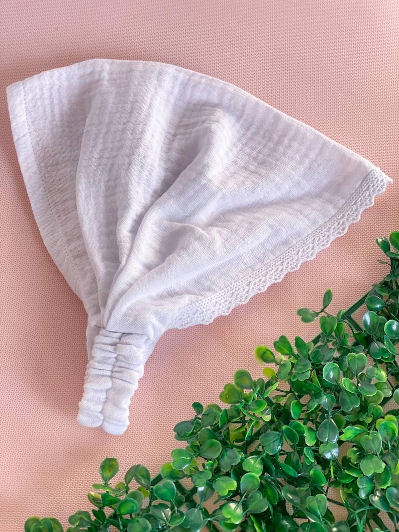 May include: A white headband with a textured fabric and a delicate lace trim. The headband is gathered at the center and appears to be made of a soft, lightweight material. The background is a light pink surface with green foliage.