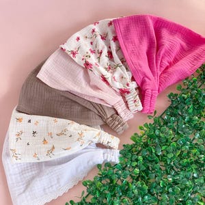 May include: An assortment of headbands in various colors and patterns. The headbands include a solid pink, a floral print on white, a light pink, a brown, and a white headband with a lace trim. The headbands are arranged on a pink surface.