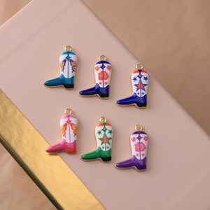 May include: Six metal charms shaped like cowboy boots, each featuring a white upper section with colorful enamel details including butterflies, roses, and stars. The boots have gold-toned edges and a small loop at the top for attachment to jewelry.