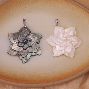 May include: Two flower-shaped pendants crafted from iridescent shell. The left pendant is dark grey with a central dark pearl, while the right pendant is white with a central white pearl. Both feature layered petals and a silver-toned metal bail.