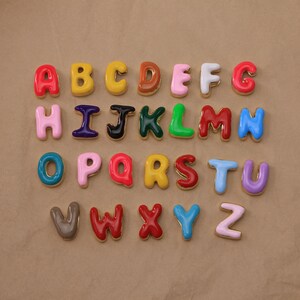 Puede incluir: Un alfabeto completo de letras tridimensionales coloridas y brillantes. Cada letra tiene un borde dorado. Las letras están dispuestas en filas y columnas sobre una superficie marrón. Las letras son de varios colores, incluyendo rojo, amarillo y azul.