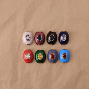 May include: Eight oval-shaped beads arranged in two rows. Each bead features a smooth, coloured enamel finish and a central gold-rimmed oval gemstone. The colours include white, maroon, black, dark blue, red, green, purple, and light blue.