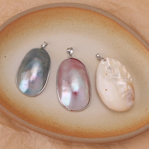 May include: Three oval-shaped pendants featuring smooth, iridescent surfaces in shades of dark grey, soft pink, and creamy white. Each pendant is encased in a simple silver-toned metal frame and includes a small metal bail at the top for hanging.