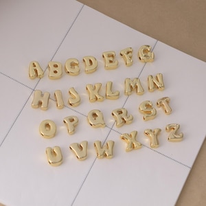 Peut inclure: Un ensemble de perles alphabet dorées, chacune avec un design arrondi en forme de bulle. Les lettres de A à Z sont affichées, parfaites pour la fabrication de bijoux ou les projets d'artisanat. Les perles sont disposées sur une surface blanche avec un motif quadrillé.