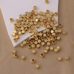 Puede incluir: Un conjunto de pequeñas cuentas hexagonales doradas dispersas sobre una superficie blanca. Las cuentas tienen un brillo metálico y un agujero en el centro, adecuado para ensartar. Estas cuentas se utilizan probablemente para la fabricación de joyas o manualidades.
