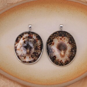 May include: Two circular pendants featuring polished brown and cream patterned seashells encased in silver-toned metal frames. Each pendant includes a small silver-toned bail at the top for attachment, showcasing natural organic textures and dark brown spots.