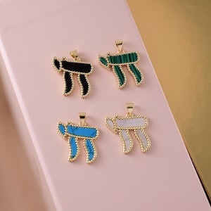 May include: Four gold-colored pendants in the shape of the Hebrew letter 'Chai'. Each pendant has a different colored inlay: black, green, turquoise, and white. The pendants are displayed on a pink surface with a gold background.