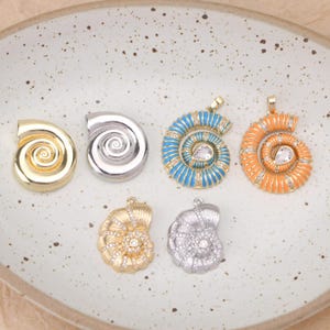 Könnte beinhalten: Sechs Nautilus-Muschel-Anhänger in zwei Reihen angeordnet. Die obere Reihe zeigt eine goldfarbene Muschel, eine silberfarbene Muschel, eine blaue Emaille-Muschel mit klaren Steinen und eine orangefarbene. Die untere Reihe enthält zwei Muscheln.