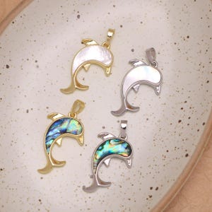 May include: Four dolphin-shaped pendants featuring inlaid shell material. Two pendants are gold-toned with white and iridescent blue-green shell, while two are silver-toned with white and iridescent blue-green shell, each with a top bail.
