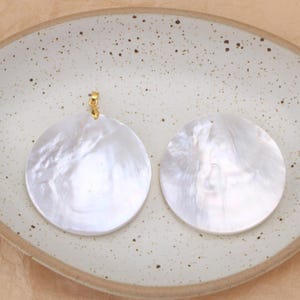 May include: Two circular, iridescent white mother-of-pearl pendants with a smooth, polished finish. One pendant features a small gold-toned metal bail attached to the top edge, while the other has a small, simple hole for suspension.
