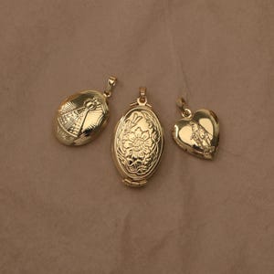 Könnte beinhalten: Drei goldfarbene Metallanhänger. Links ein ovales Medaillon mit Strahlenmuster und einer Figur. In der Mitte ein ovales Medaillon mit geprägtem Blumenmuster. Rechts ein herzförmiges Medaillon mit einer eingravierten religiösen Figur.