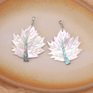 Op de afbeelding: Twee bladvormige hangers gemaakt van iriserend wit parelmoer met een centrale steelinleg van kleurrijke abalone schelp. Elke hanger is voorzien van een zilverkleurig metalen oogje aan de bovenkant voor bevestiging en heeft een gedetailleerd nerfpatroon.