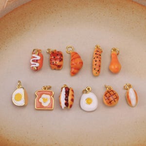 Può includere: Una collezione di undici piccoli ciondoli in metallo a tema alimentare con dettagli in smalto. I ciondoli includono rappresentazioni in miniatura di un hot dog, sushi, croissant, baguette, pera, uovo a fette, toast con uovo, sandwich, uovo al tegamino, waffle e panino.