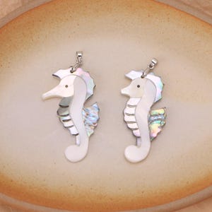 Peut inclure: Deux pendentifs en forme d'hippocampe fabriqués en nacre irisée et en coquillage blanc. Chaque hippocampe présente un corps blanc lisse, un œil sombre et une nageoire texturée aux tons nacrés chatoyants et multicolores, avec une bélière en métal argenté.