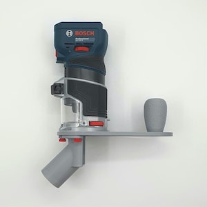 Könnte beinhalten: Ein Bosch Professional Elektrowerkzeug, mit blauem und schwarzem Gehäuse und roten Akzenten. Montiert auf einer grauen Plattform mit Griff und Staubabsauganschluss, für Holzbearbeitung und Bauarbeiten.