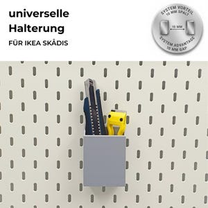 Könnte beinhalten: Grauer, rechteckiger Aufbewahrungsbehälter mit einem Cuttermesser, einem blauen Cutter und einem gelben Werkzeug. Der Behälter ist an einer weißen Lochwand befestigt. Der Text auf dem Bild ist auf Deutsch.