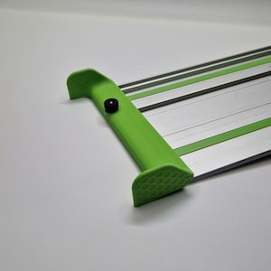 Könnte beinhalten: Ein Papierschneider mit einem leuchtend grünen Kunststoffgriff, der an der Seite ein strukturiertes Wabenmuster und einen kleinen schwarzen runden Knopf aufweist. Die Basis besteht aus einer langen, flachen silbernen Metallfläche.