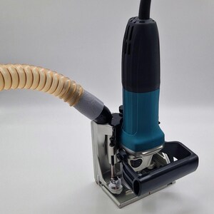 Puede incluir: Una herramienta eléctrica de color verde azulado y negro con una carcasa de motor vertical, un mango negro y una base de metal plateado. Una manguera flexible de color beige translúcido está unida al lateral para extraer polvo hacia la izquierda.