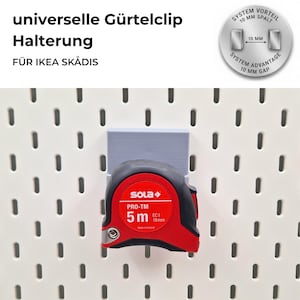 Puede incluir: Un flexómetro rojo y negro, marcado "SOLA PRO-TM 5 m", está sujeto a un soporte gris sobre una pared perforada blanca. La imagen incluye texto en alemán. El flexómetro mide 19 mm de ancho.