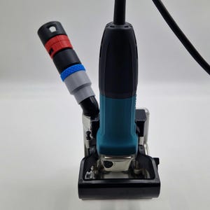Könnte beinhalten: Ein blaugrünes und schwarzes Elektrowerkzeug mit vertikalem Griff und einem Stromkabel, das oben herausragt. Ein abgewinkelter Aufsatz mit roten, schwarzen, blauen und grauen Segmenten ist seitlich angebracht. Die Basis ist ein schwarz-silberner Metallrahmen.