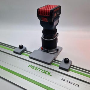 Puede incluir: Una fresadora inalámbrica con una parte superior roja y negra, un cuerpo azul y una base negra está montada en un sistema de rieles gris. La pista es plateada con detalles en verde y la marca "FESTOOL" está impresa.