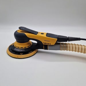 Könnte beinhalten: Ein gelb-schwarzer elektrischer Exzenterschleifer Mirka DEROS mit rundem Schleifteller, einem schwarzen Bedienhebel mit der Aufschrift 5.0 und einem flexiblen, durchscheinenden, gewellten Vakuumschlauch zur Staubabsaugung am hinteren Ende.