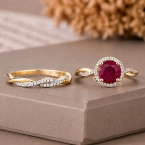 May include: Two gold rings. One ring features a large, round, red gemstone surrounded by small white stones. The other ring is a gold band with a twisted design and small white stones.