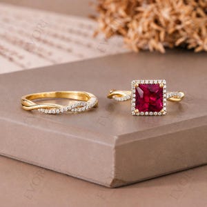 Puede incluir: Dos anillos de oro. Uno presenta una gema roja cuadrada rodeada por un halo de piedras más pequeñas. El segundo es una banda de oro con un diseño retorcido y pequeñas piedras transparentes. Ambos anillos están sobre una superficie marrón.