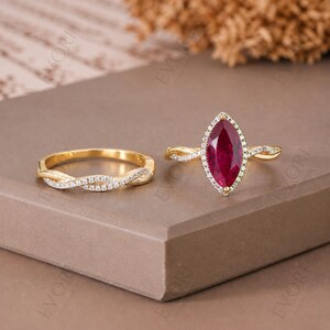 May include: Two gold rings. One ring features a marquise-cut ruby surrounded by small diamonds, set in a twisted band. The second ring is a gold band with a twisted design and small diamonds. The rings are displayed on a brown surface.