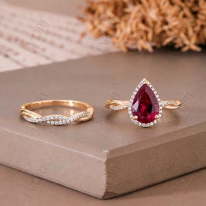 Op de afbeelding: Een tweedelige sieradenset met een gouden ring met een gedraaide band en een gouden verlovingsring. De verlovingsring heeft een peervormige, dieprode edelsteen omringd door kleine, heldere stenen. De ringen staan tegen een neutrale achtergrond.