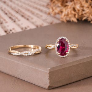 Op de afbeelding: Een set van twee gouden ringen. Eén ring heeft een grote ovale, dieprode edelsteen omringd door kleine witte stenen. De tweede ring is een gouden band met een gedraaid ontwerp en kleine witte stenen. Beide ringen liggen op een bruin oppervlak.