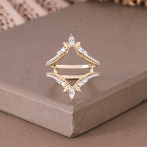 Può includere: Un anello di protezione dorato con un design a forma di foglia. L'anello di protezione presenta una serie di piccole pietre preziose trasparenti e una pietra centrale a forma di marquise. Progettato per anelli di fidanzamento solitari.