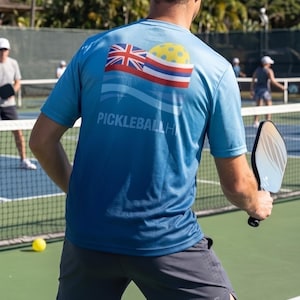 Pu&ograve; includere: Una maglietta sportiva blu a maniche corte con una grafica sul retro che mostra la bandiera dello stato delle Hawaii, una pallina da pickleball gialla e il testo PICKLEBALLHI. La maglietta &egrave; abbinata a pantaloncini sportivi grigio scuro e una racchetta nera.