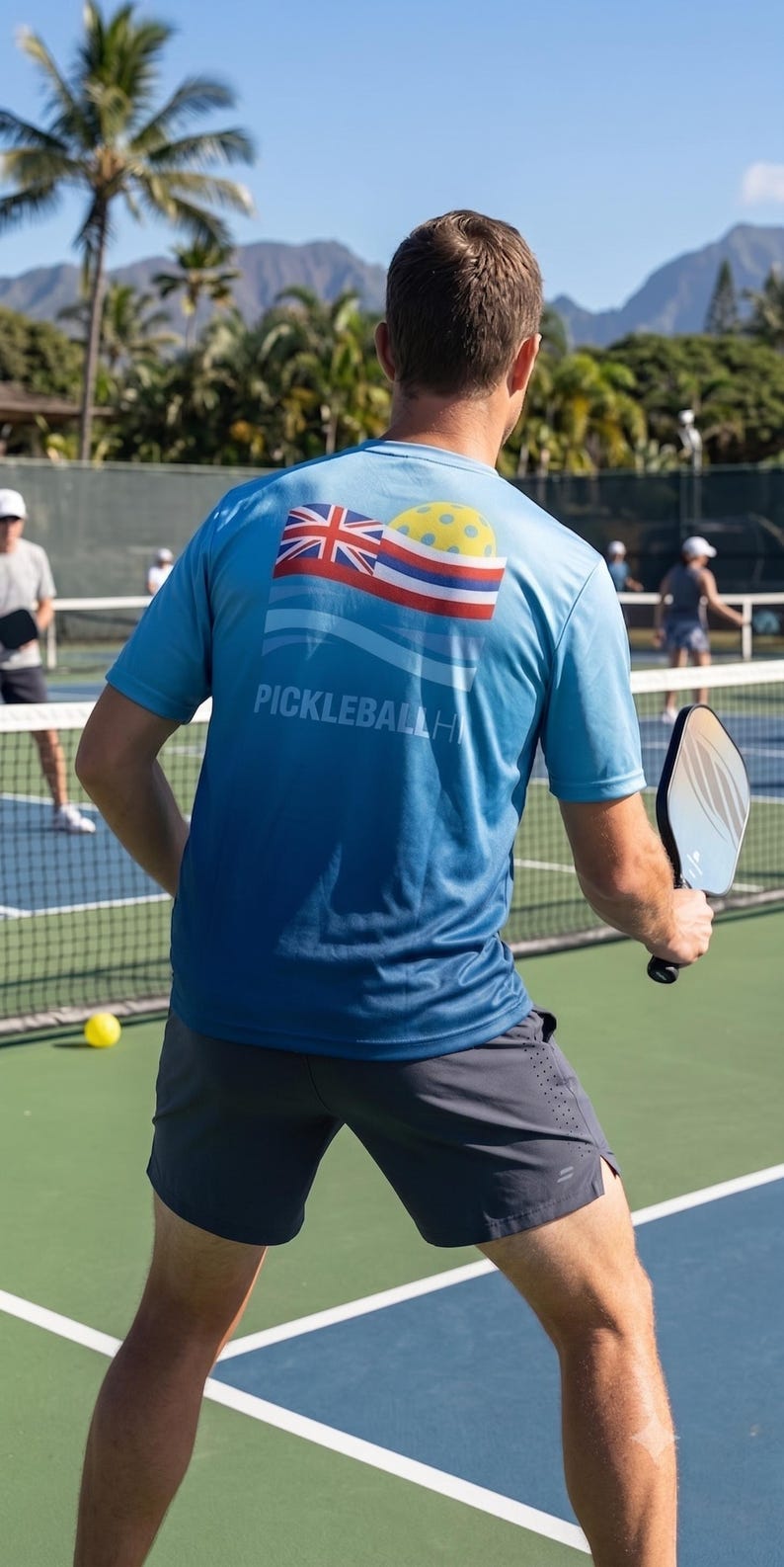 Pu&ograve; includere: Una maglietta sportiva blu a maniche corte con una grafica sul retro che mostra la bandiera dello stato delle Hawaii, una pallina da pickleball gialla e il testo PICKLEBALLHI. La maglietta &egrave; abbinata a pantaloncini sportivi grigio scuro e una racchetta nera.