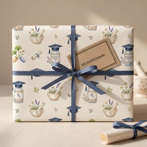 May include: Wrapped gift with a pharmacy-themed pattern featuring mortar and pestle, graduation caps, and prescription bottles. A navy blue ribbon and a tag that reads "MICHAELBOSTUDIO" adorn the gift. A rolled-up diploma is visible.